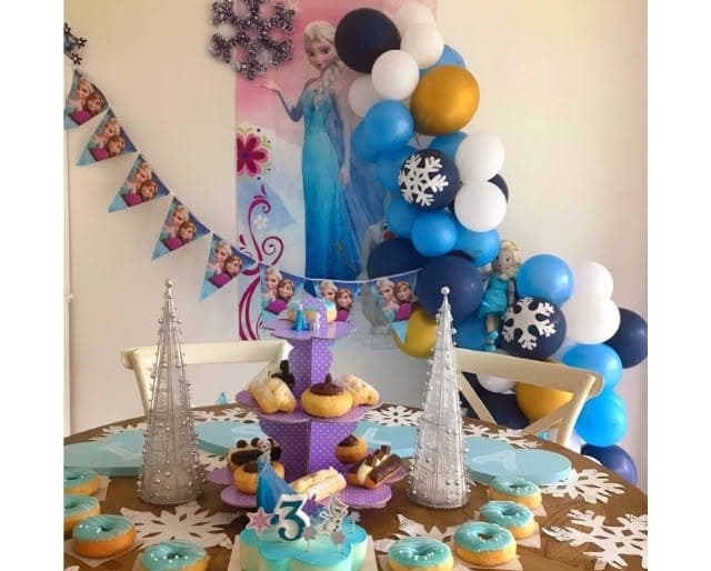 Frozen-themed third birthday party table with Elsa poster, balloon arch, blue donuts, and cake.