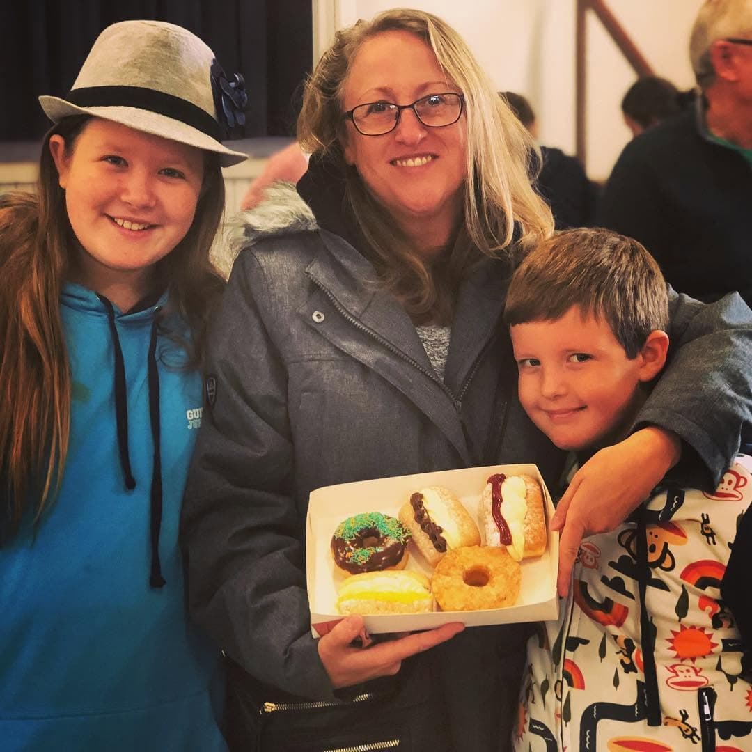 Smiling woman and two children holding a white box filled with five assorted donuts.