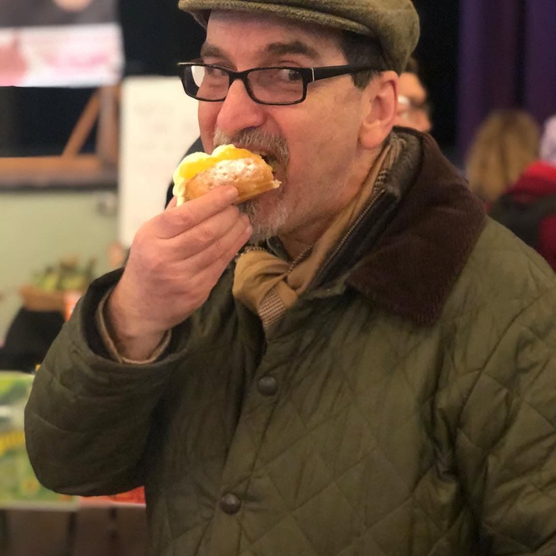 Man in a flat cap and glasses takes a bite of a cream-filled pastry.