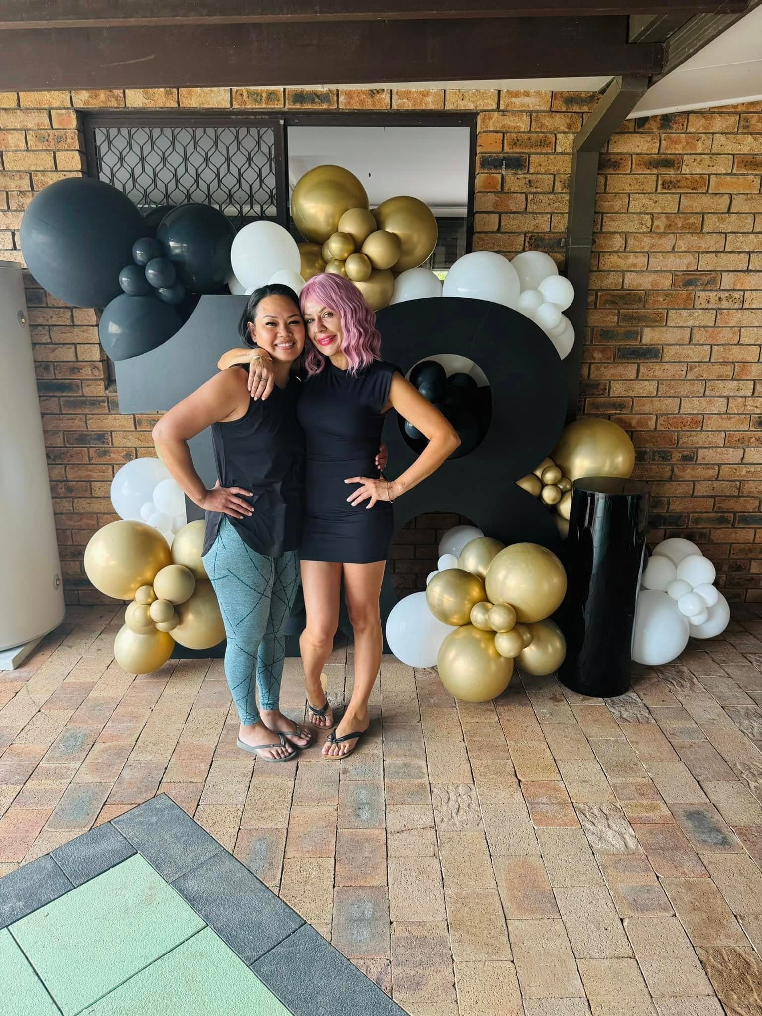Two women pose before a large 18 display decorated with black, white, and gold balloons.