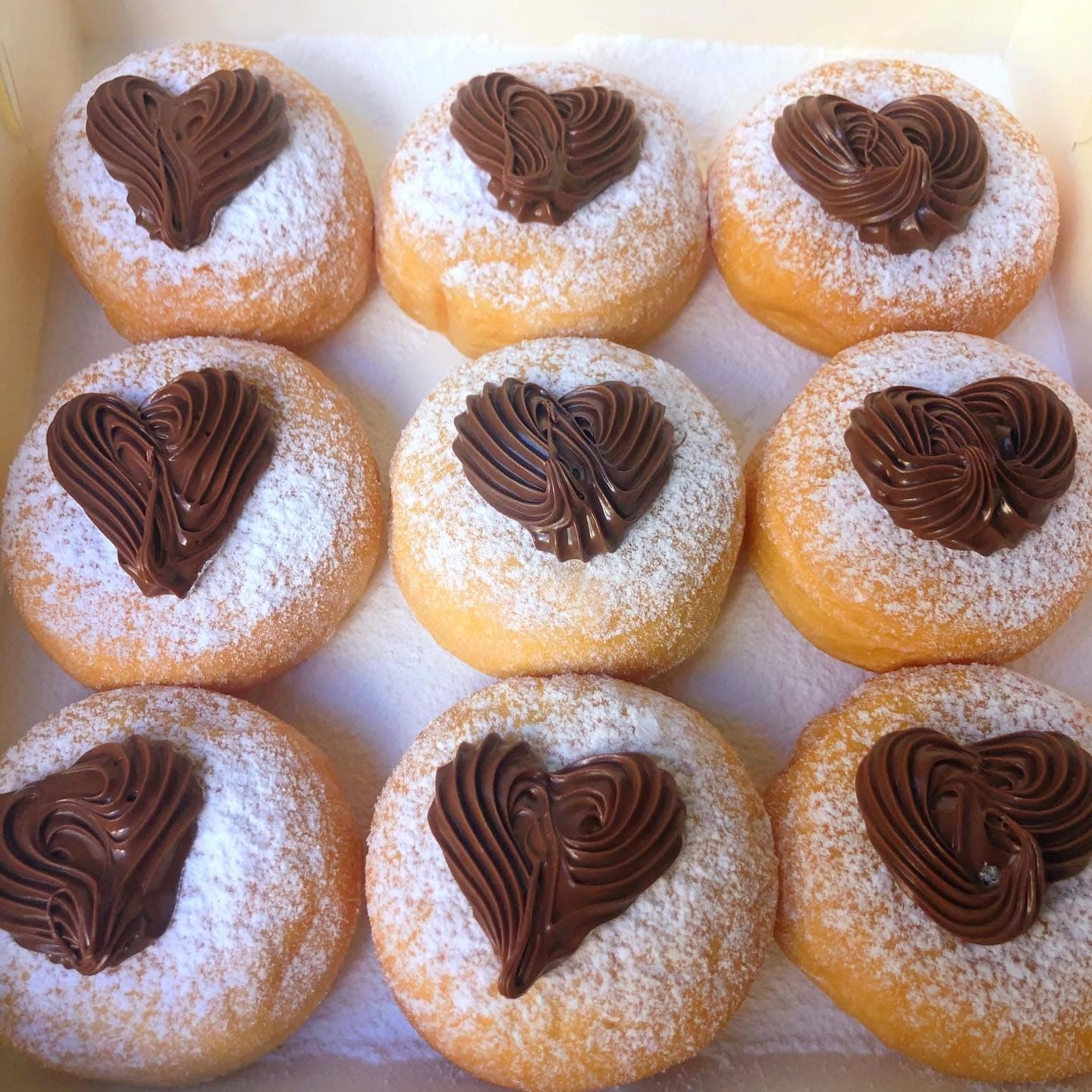 Nine powdered sugar donuts in a box, each topped with a heart-shaped chocolate frosting swirl.