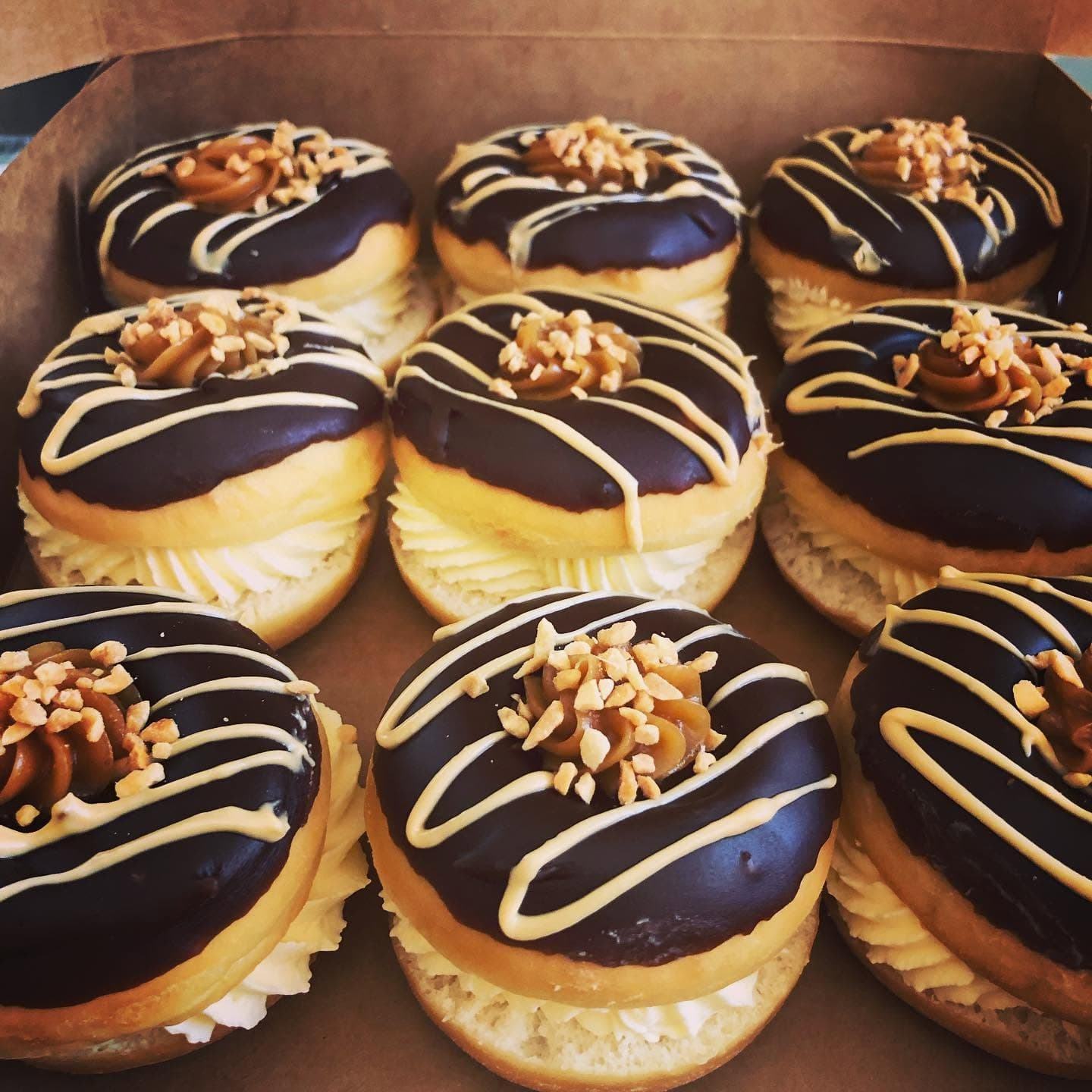 Nine chocolate-glazed donuts with cream filling, caramel drizzle, and crushed nuts in a box.