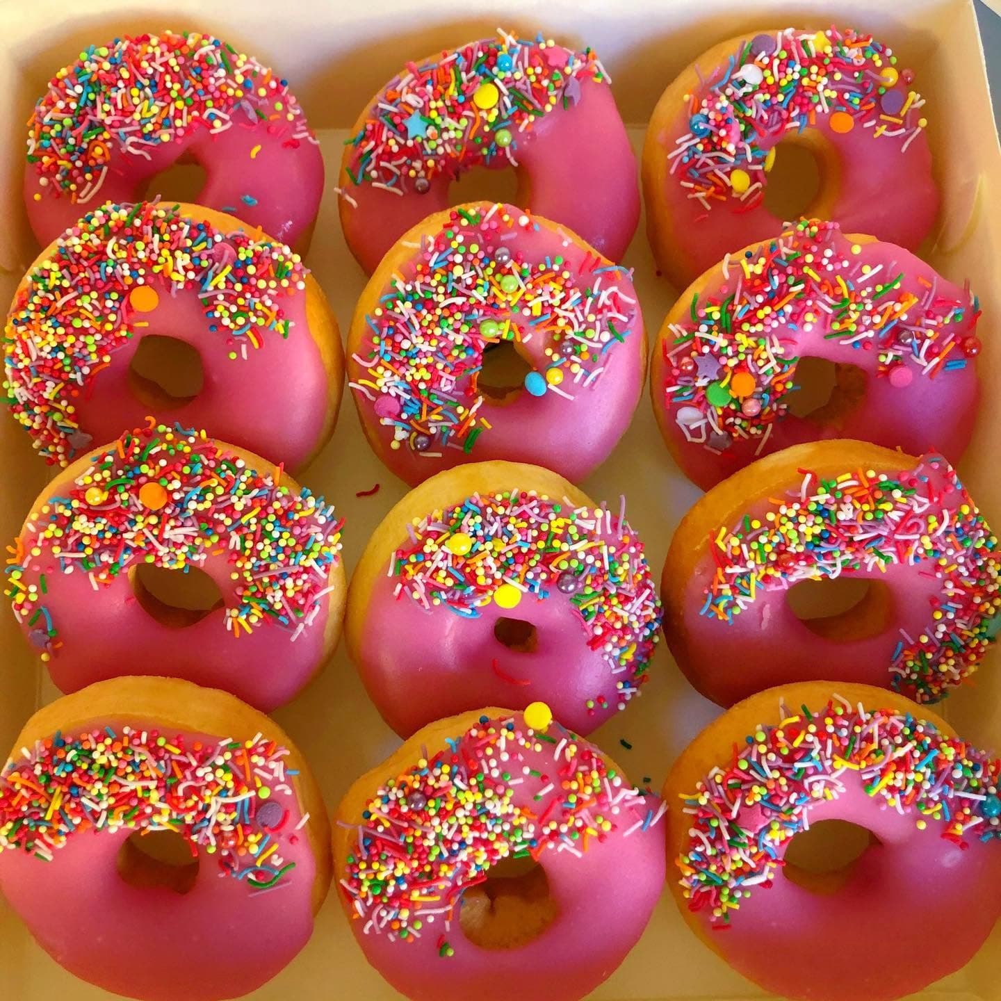 Twelve donuts with bright pink frosting and colorful rainbow sprinkles arranged in a white box.