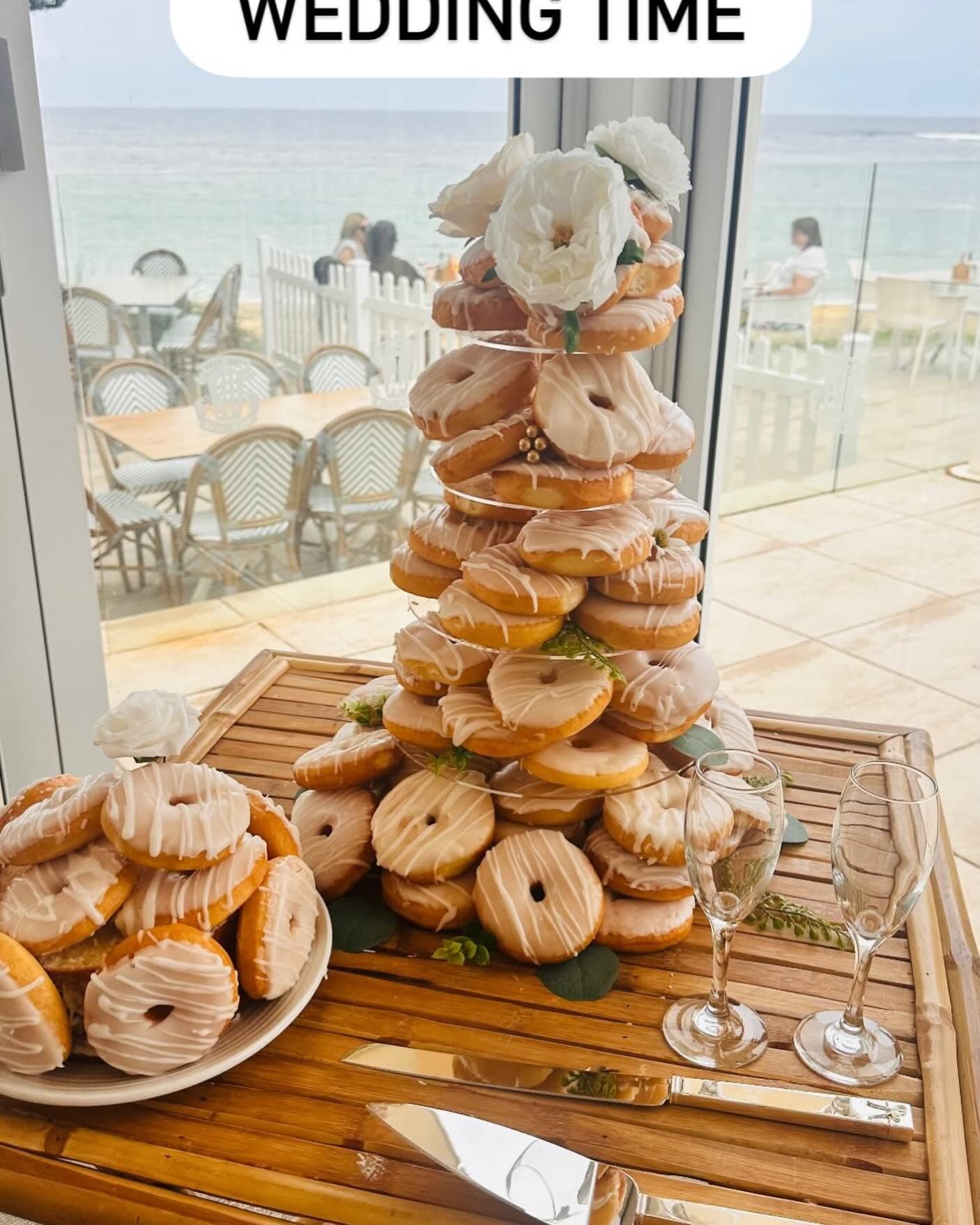 Tiered glazed donut tower with white flowers and champagne glasses on a seaside table.